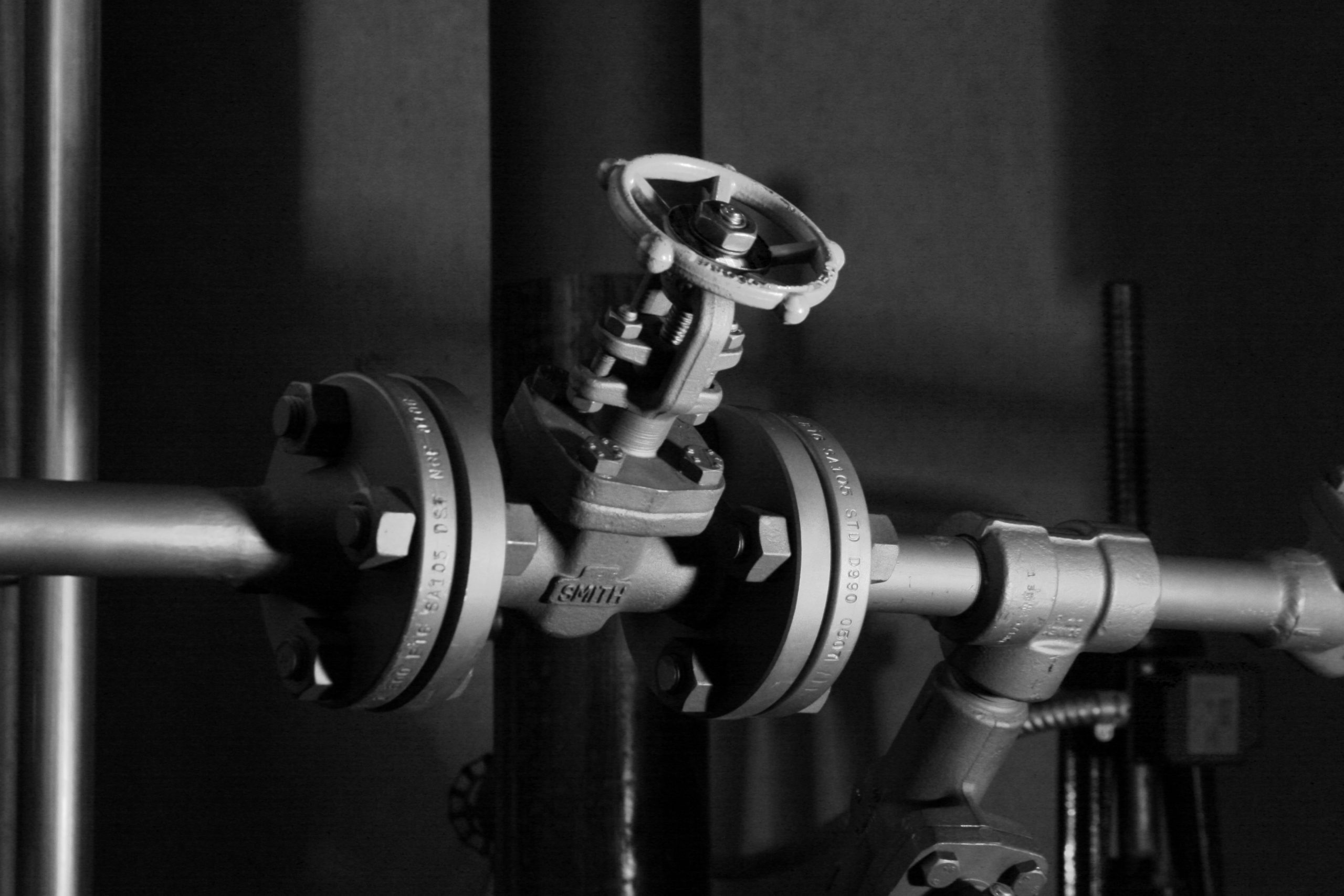 A black and white photo of a metal valve with a handwheel, connected to horizontal pipes by large bolts. Shadows and lighting emphasize the industrial setting, reflecting a half century of reputation in industrial design.