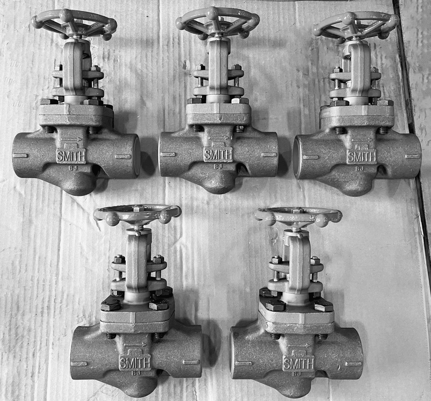 Five metal gate valves, labeled SMITH and featuring handwheels for manual operation, are arranged in a V shape on a cardboard surface. The black-and-white image highlights key differences often specified by ISO standards in valve design.
