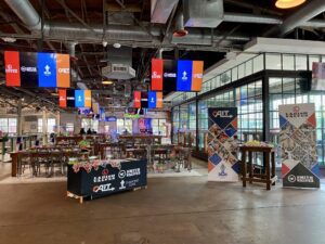 Spacious indoor event venue with high ceilings, colorful hanging screens, long wooden tables, branded covers, and standing banners showcasing Valve World Americas Expo. Glass walls on one side create a bright setting for Customer Appreciation gatherings.